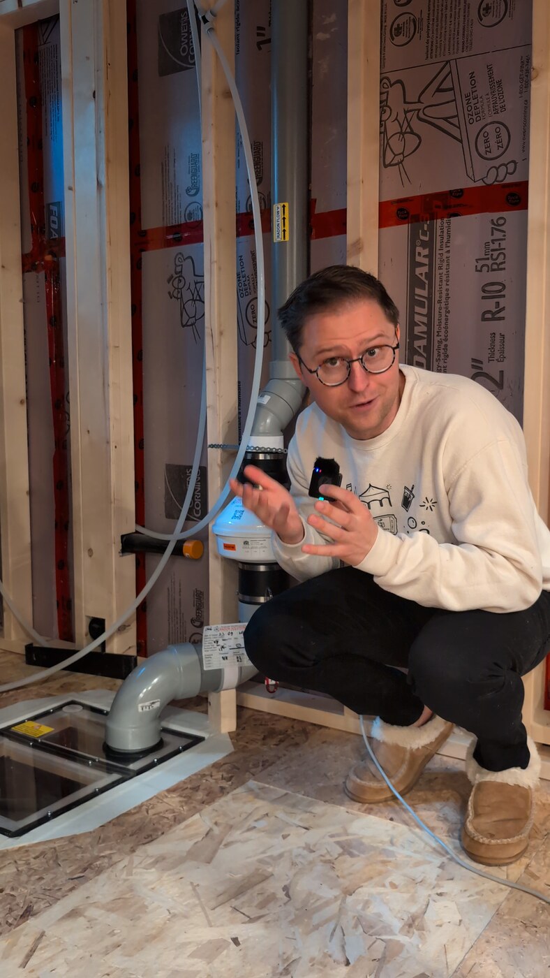 Troy crouched in his unfinished basement next to the finished radon mitigation system: a white fan on a grey pipe running down into the slab.