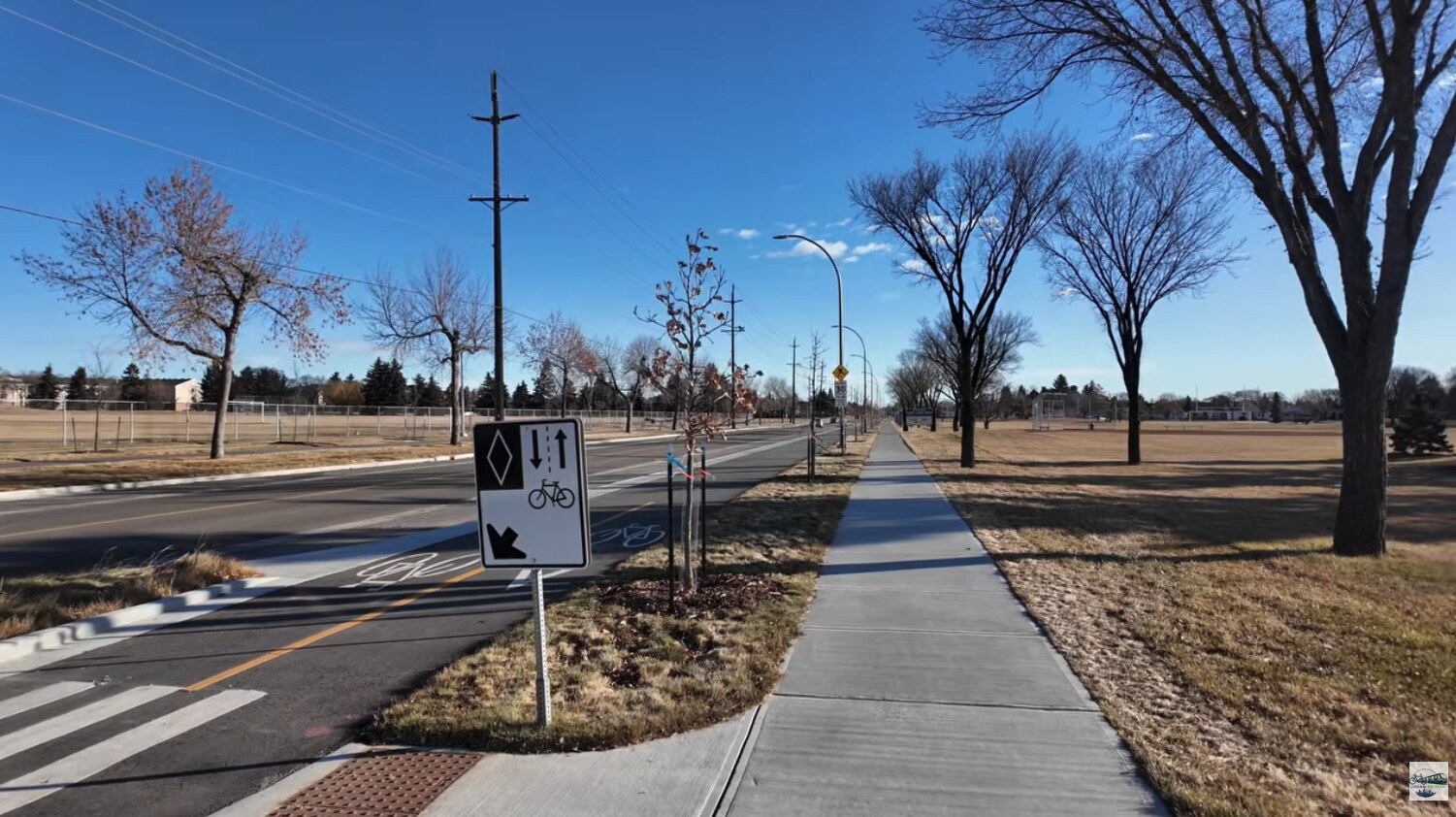 132 Avenue in Edmonton: a painted bike lane beside the road, a boulevard with young trees, and a sidewalk running past a park on a sunny day.